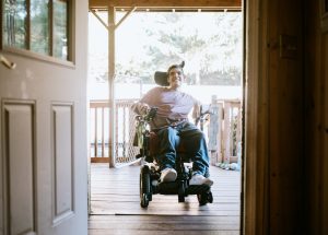 A cheerful young adult man with cerebral palsy drives up a wheelchair ramp to enter his home.  Horizontal image with copy space.